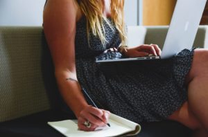 Une femme assis sur un canapé regarde son ordinateur sur ses genaoux tout en écrivant à la main sur un carnet de note